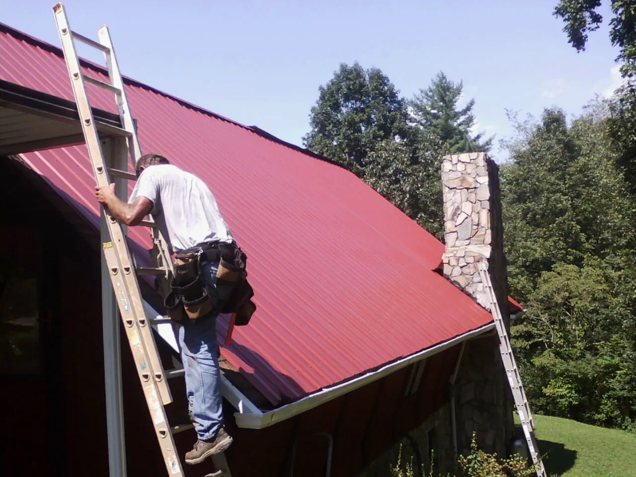 Roofer on a ladder beside a red metal roof and stone chimney — Soffit & Fascia Repair by Roofing Brothers in Kalama